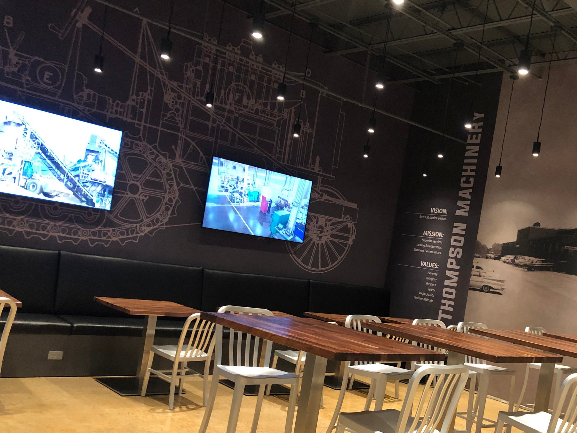 Modern dining area with wooden tables and metal chairs against a wall with large screens and branding.