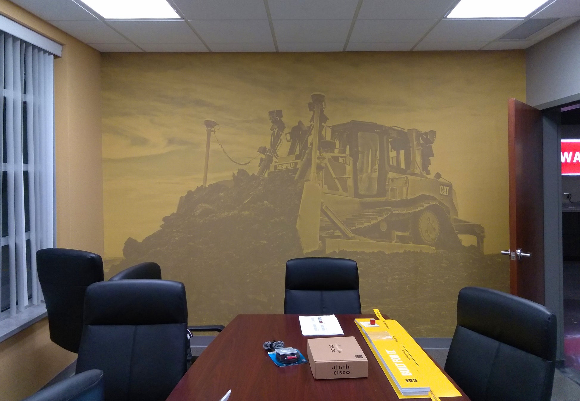 A large wall mural in an office setting depicting construction equipment, applied to the wall behind a meeting room table.